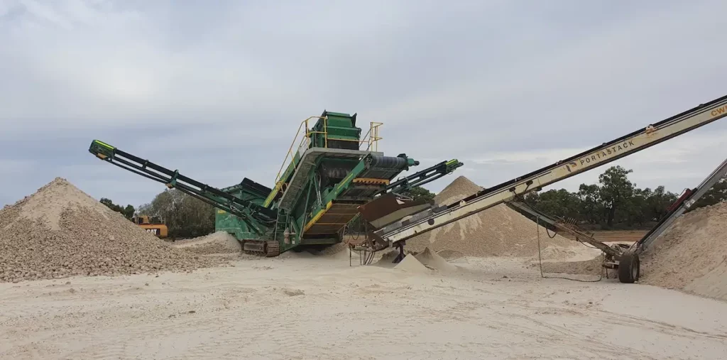 A large green sand screening machine operates outdoors, sorting and moving sand onto tall piles with conveyor belts under a cloudy sky. Trees line the background of the industrial site.