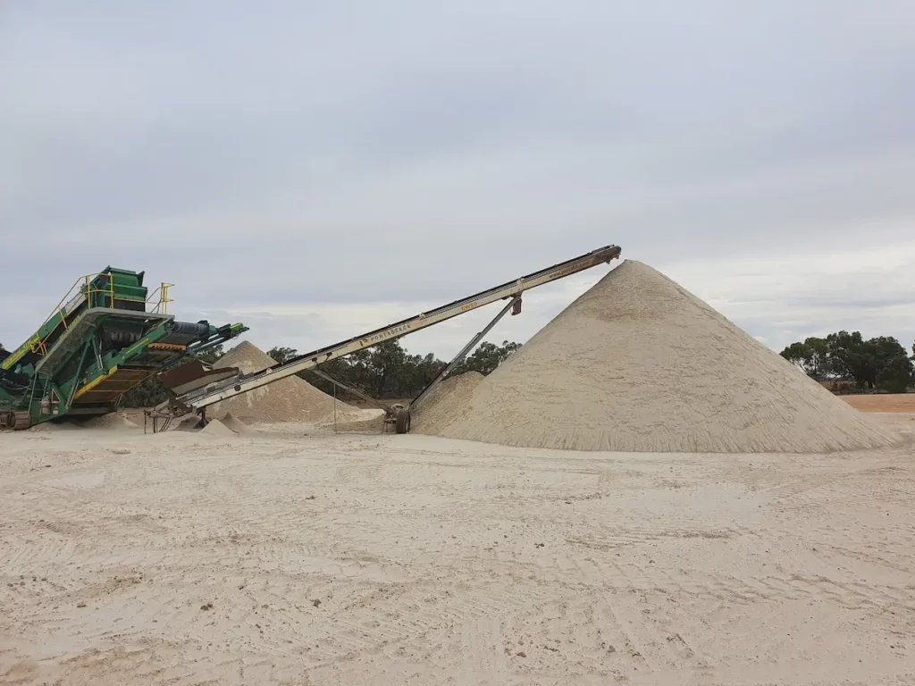 A conveyor belt deposits sand onto a large conical pile at a sand quarry, with machinery and smaller sand mounds visible in the background under a cloudy sky.