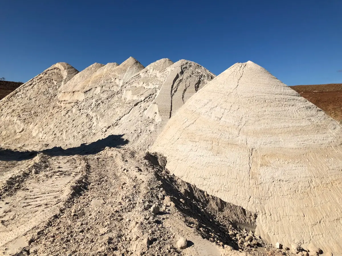 Large conical piles of white sand or mineral material are stacked under a clear blue sky in a barren, open landscape. Tire tracks are visible in the dirt in front of the piles.