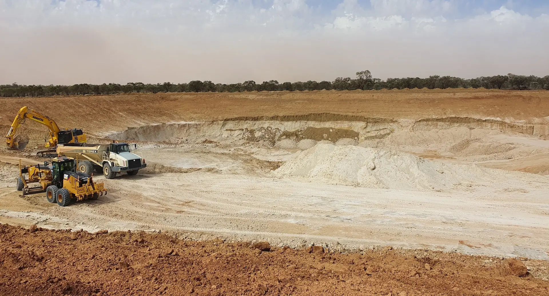 A wide, open mining site with an excavator, a dump truck, and a grader working on sandy, reddish-brown earth under a partly cloudy sky, with sparse trees lining the distant horizon.