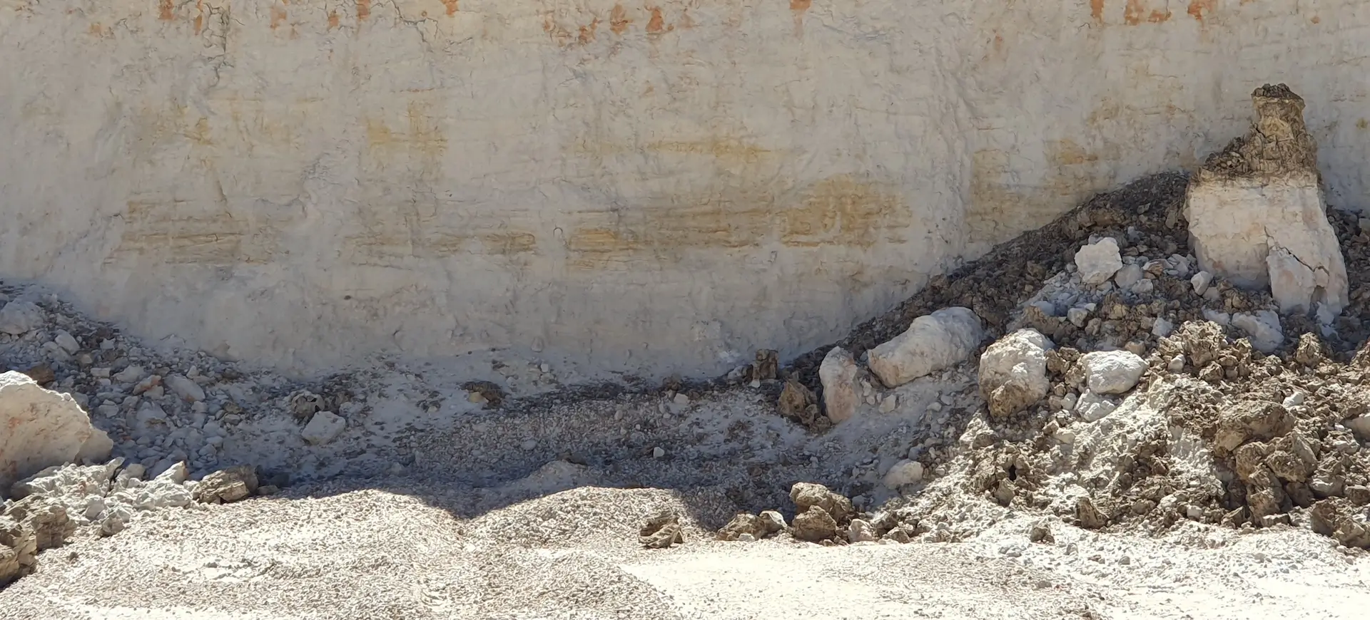 A sandy excavation site with a vertical dirt wall, piles of loose rubble, rocks, and gravel scattered at the base. The scene appears sunlit with shadows cast along the ground and debris.